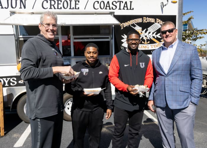 Group of men in front of a food truck with food in hand smiling at the camera