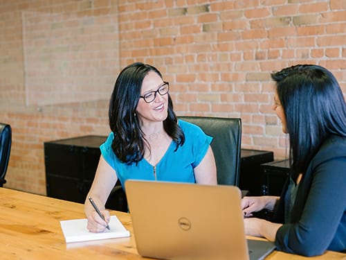 two women sitting at a table talking, one with a laptop and one taking notes